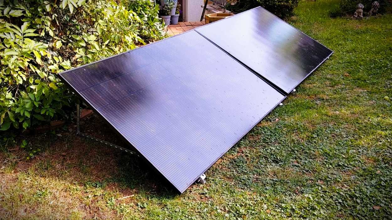 Two black solar panels positioned on a garden lawn near bushes, angled to capture sunlight.