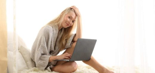 Woman sitting on bed with a laptop, smiling while working in a cozy sweater.